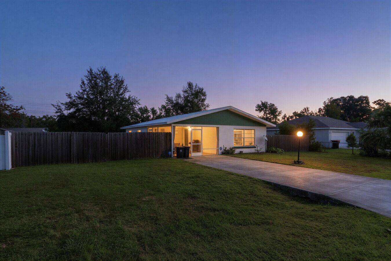 Une maison de plain-pied avec un porche à l’avant, un jardin clôturé et un éclairage extérieur suffisant au crépuscule.