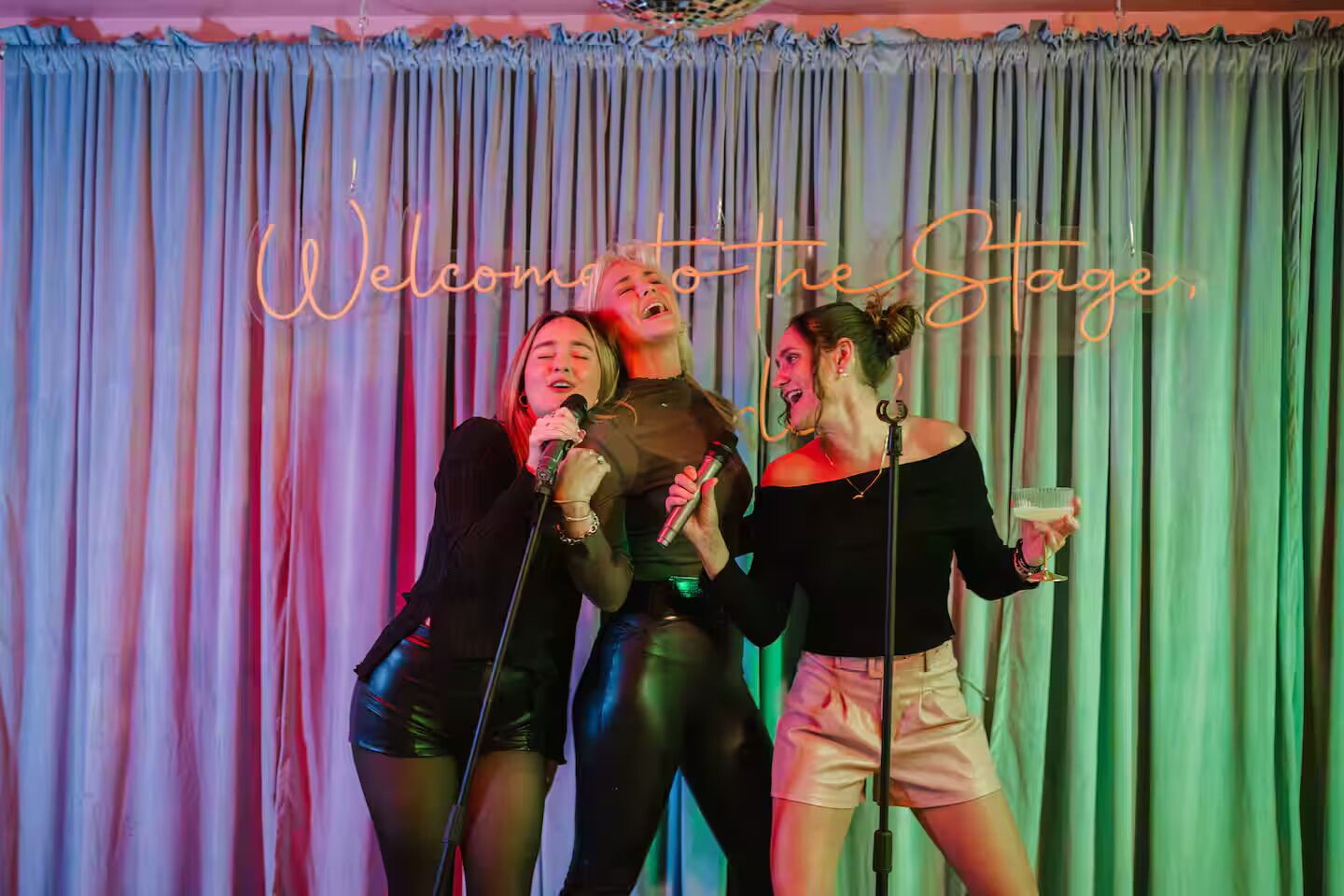 Tres personas en el escenario con micrófonos, un letrero de neón que dice "Welcome to the Stage" y un telón de fondo.