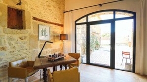 Une pièce avec un mur en pierre, une table en bois sur laquelle se trouve un jeu d’échecs, deux chaises, une lampe et une grande porte vitrée donnant sur l’extérieur.