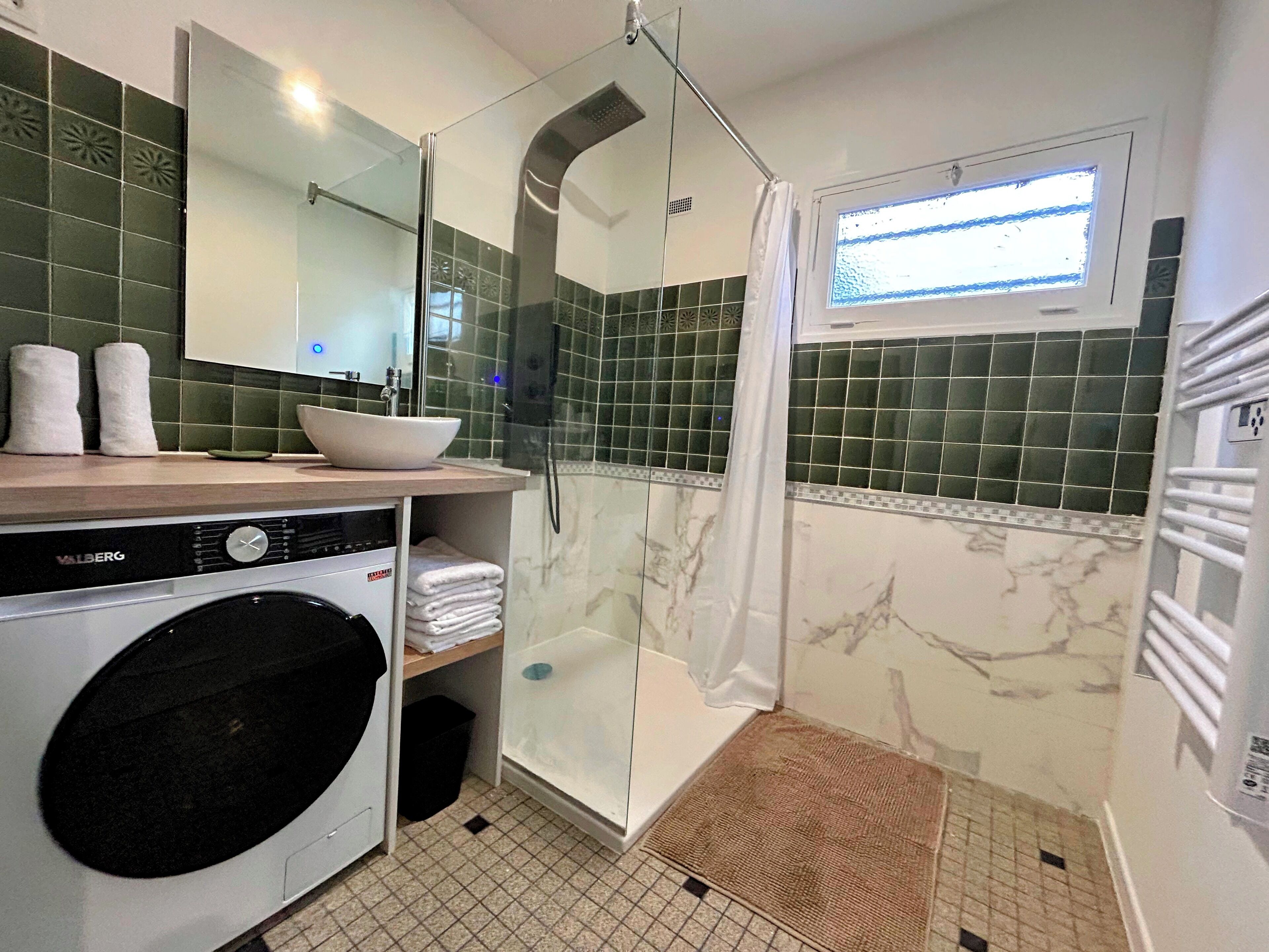 Une salle de bain compacte dotée d’une douche à parois de verre, d’un lavabo en forme de cuvette, d’un lave-linge et d’une fenêtre laissant entrer la lumière naturelle.
