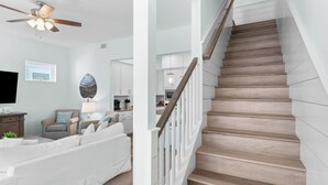 A modern living room with a sofa, a ceiling fan, and a kitchen in the background. - Constant Getaway by Avantstay 30A Coastal Retreat Outdoor Lounge, Golf Cart, Bikes + Near Beach (Santa Rosa Beach)