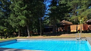 A swimming pool with a wooden deck, surrounded by trees and a house in the background.