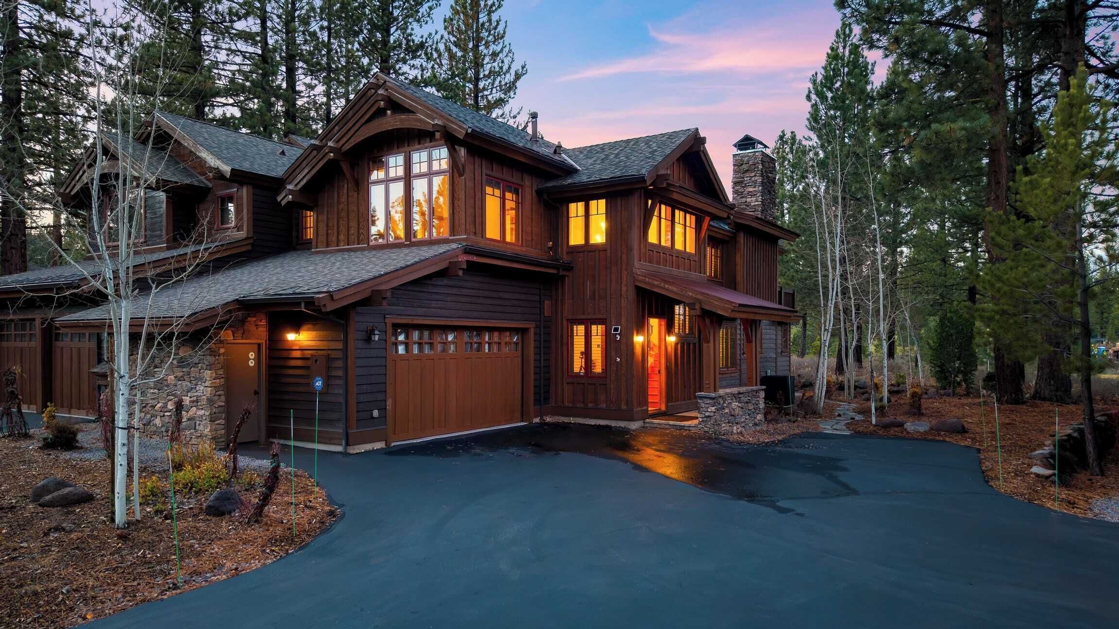 A large, two-story log cabin with a stone chimney, multiple windows, and a driveway leading to a garage.