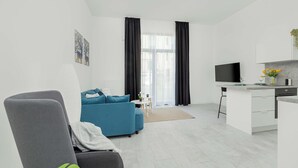 Un soggiorno moderno con un divano blu, una poltrona grigia, un piccolo tavolo da pranzo e un angolo cucina con TV e una pianta.