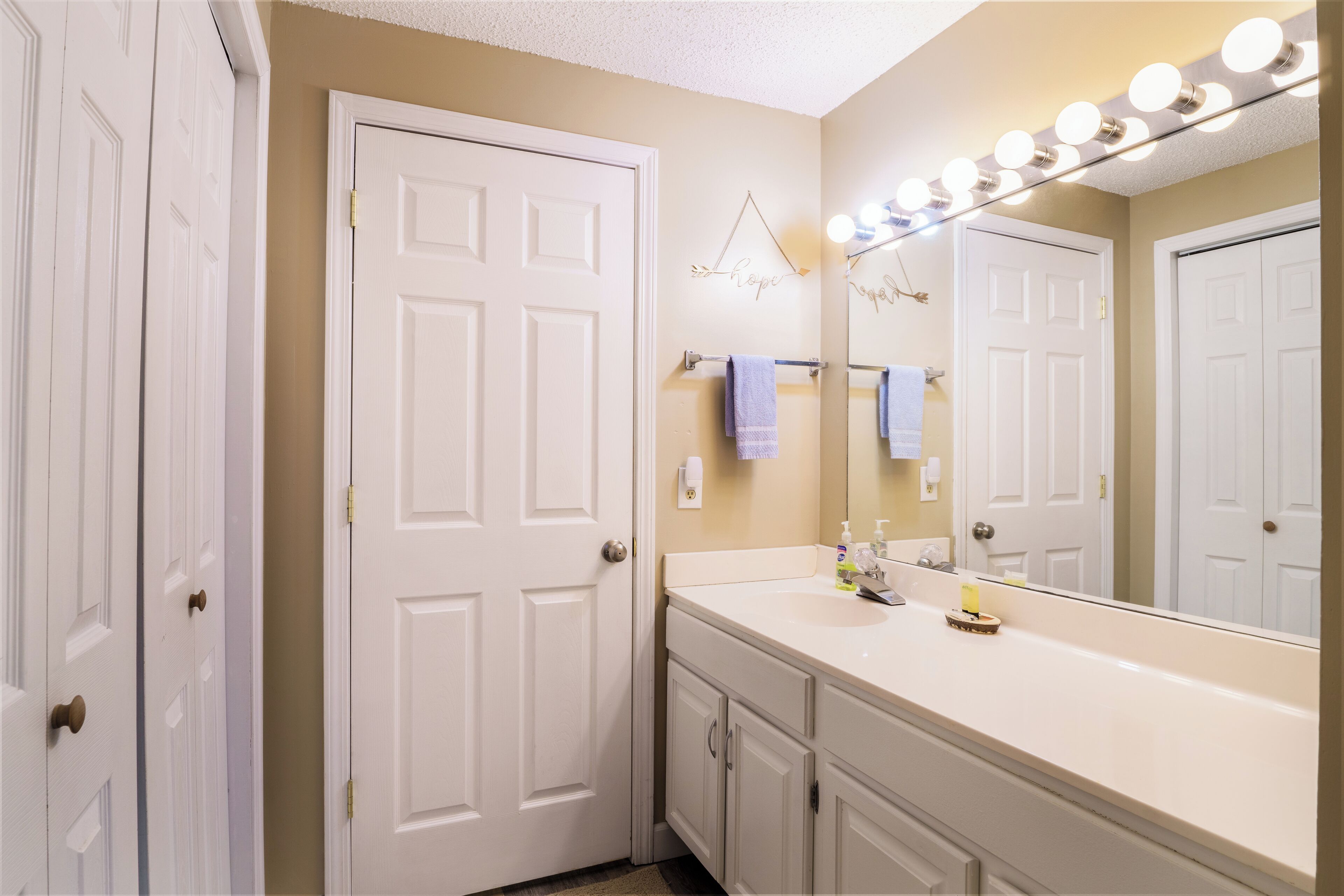 A bathroom with a white vanity, a large mirror with lights, and a towel rack.