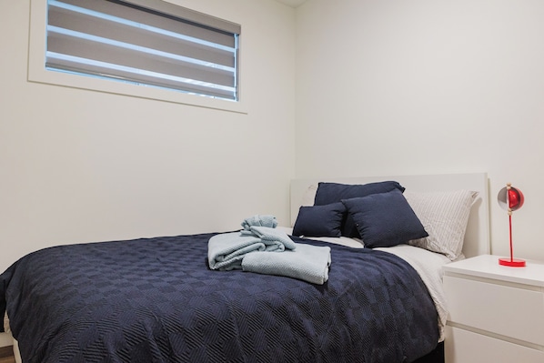 A neatly made bed with a dark blue quilt, a white bedside table with a red lamp, and a window with blinds.