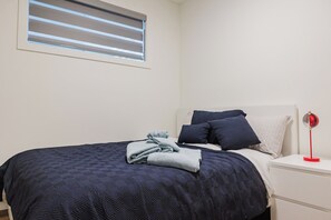 A neatly made bed with a dark blue quilt, a white bedside table with a red lamp, and a window with blinds. - Runway Retreat -2bed -1bath Basement suite 10 mins to YEG Airport (Leduc)