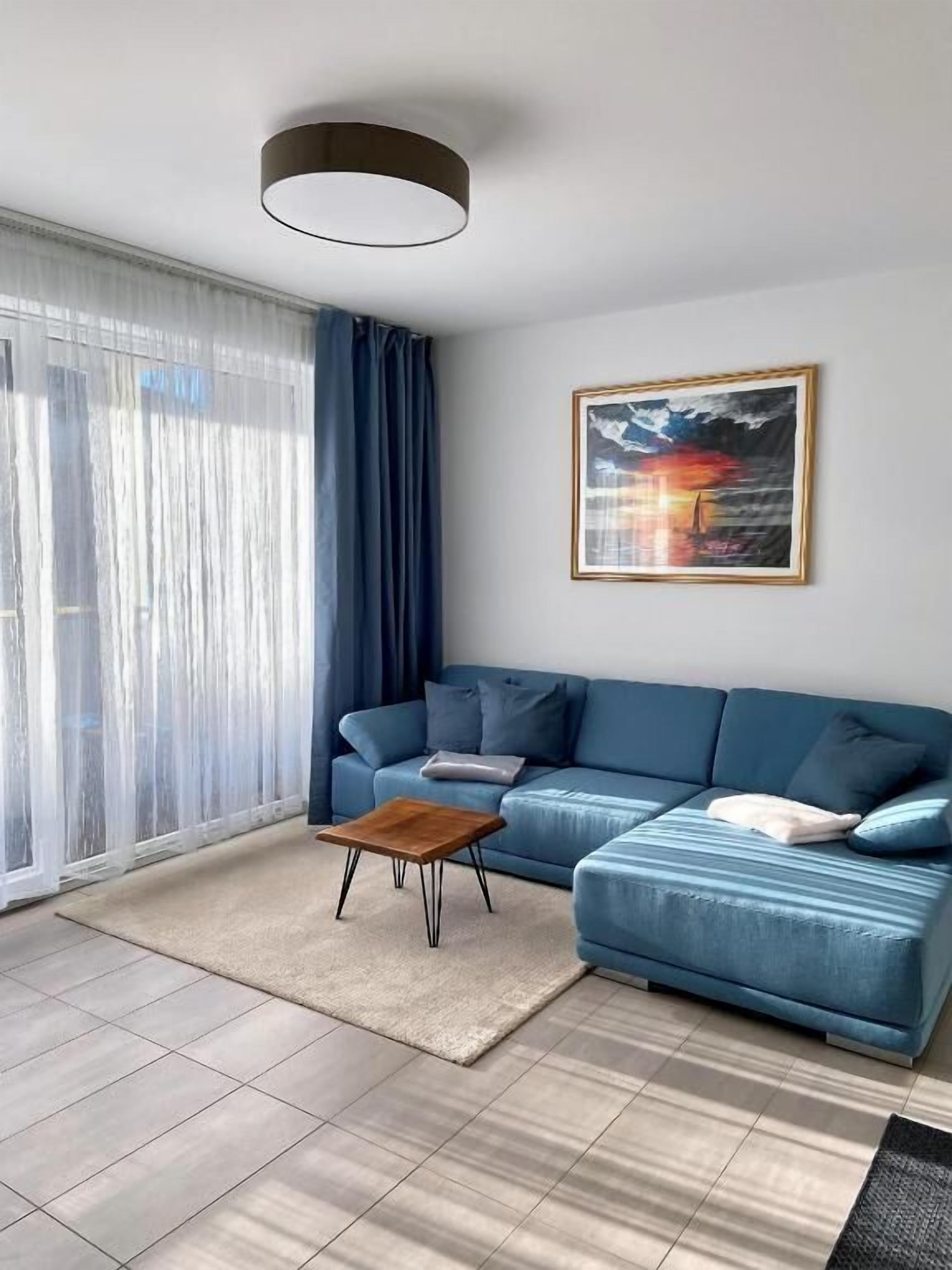 Ett modernt vardagsrum med en blå soffa, ett träbord och ett stort fönster med tunna, genomskinliga gardiner.