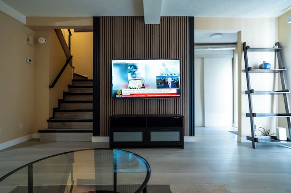 A modern living room with a flat-screen TV mounted on a wooden panel wall, a glass coffee table, a black entertainment unit, and a wooden staircase.