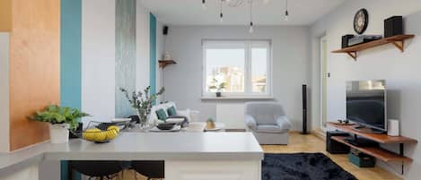 A modern living space with a kitchen island, dining area, and seating. There is a large window providing natural light and a view of the city.