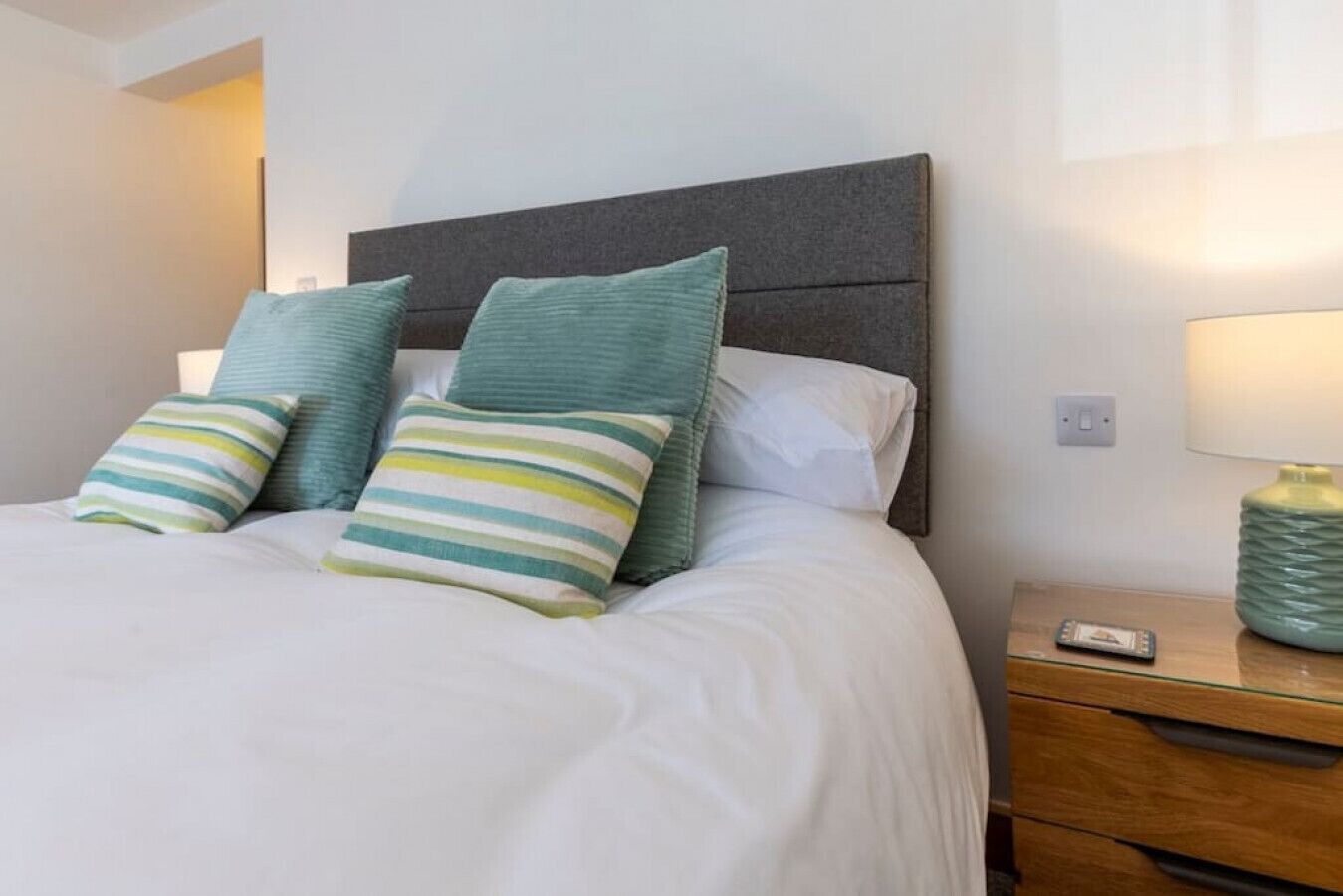 A neatly made bed with white bedding and striped pillows, a dark headboard, and a bedside table with a lamp and a decorative vase.