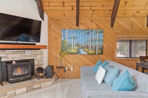 Ein gemütliches Wohnzimmer mit Kamin aus Stein, einem flachbildfernseher darüber und einem Gemälde einer Waldszene an der Wand.
