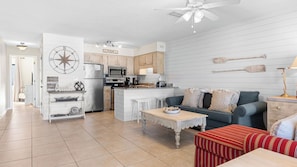 A modern kitchen with a central island, a dining area with a red-striped ottoman, and a living room with a gray sofa. - Steps to the Beach Pool, Tennis, & Putting Green Southwinds H12 by Avantstay (Panama City Beach)