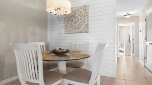 A dining area with a round glass table, white chairs, a chandelier, and a decorative wall piece. - Steps to the Beach Pool, Tennis, & Putting Green Southwinds H12 by Avantstay (Panama City Beach)