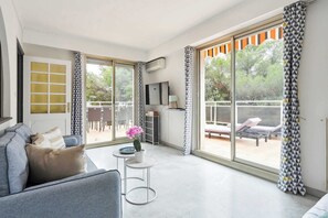Ein geräumiges Wohnzimmer mit einem grauen Sofa, einem kleinen runden Tisch mit einer Topfpflanze und einer großen Schiebeglastür, die zu einem Außenbereich mit Pool und Liegestühlen führt.