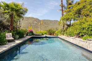A clear swimming pool with a stone border, surrounded by lush greenery and a chair.