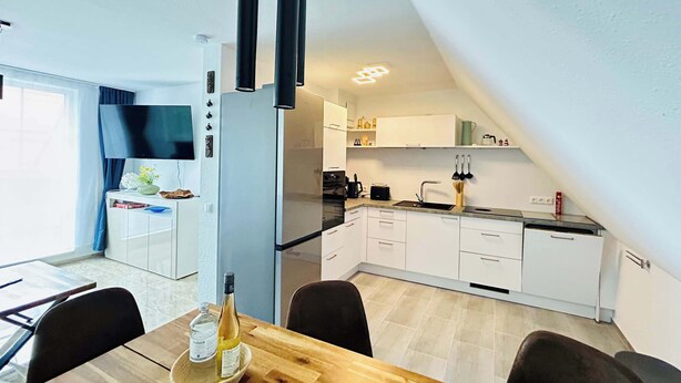 A modern kitchen with white cabinets, a black countertop, and a dining area with a wooden table and chairs. - FeWo II Graal-Müritz Sobierajczyk (Graal-Müritz)