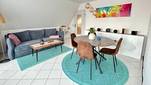Un salón moderno con un sofá gris, una mesa de centro de madera y una mesa de comedor redonda con sillas marrones. En la pared, un cuadro abstracto y colorido, y un mueble blanco con objetos decorativos.