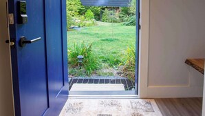 Een blauwe deur die uitkomt op een tuin met een zonnepaneel, een klein plankje en een kamerkleed met een patroon.