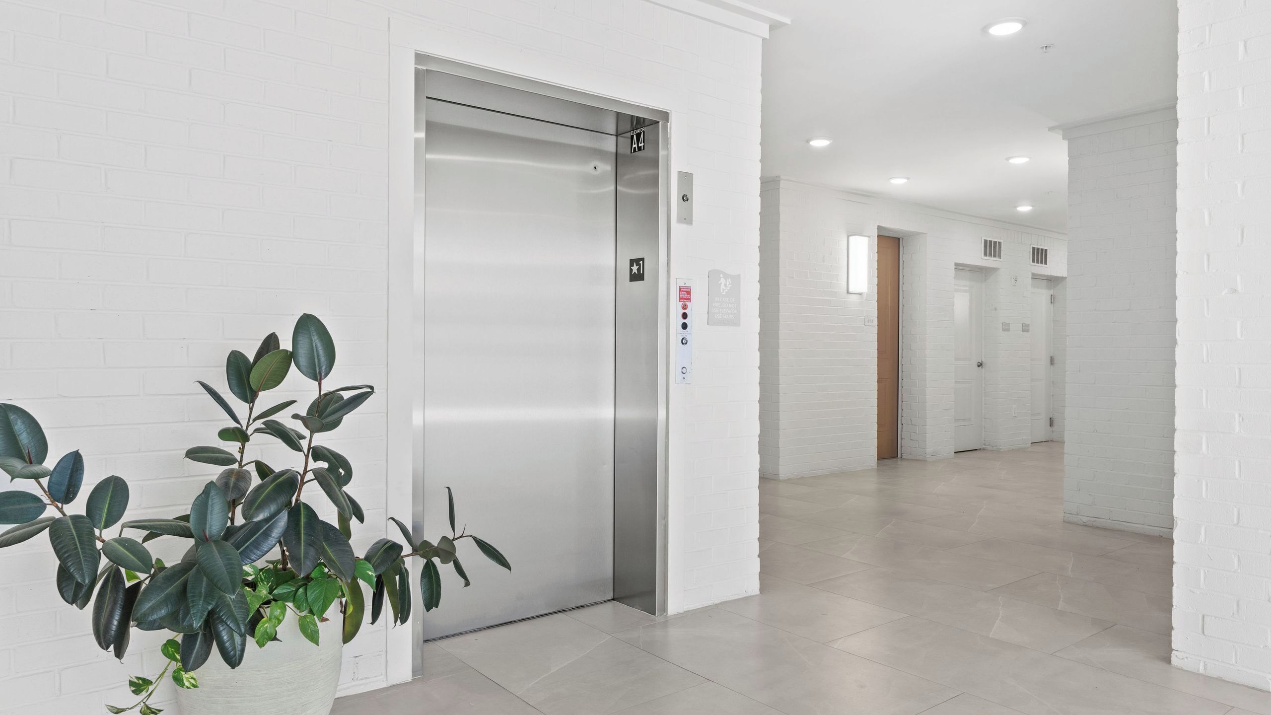 A modern elevator with a control panel, a potted plant, and a clean, white-walled hallway leading to another elevator.
