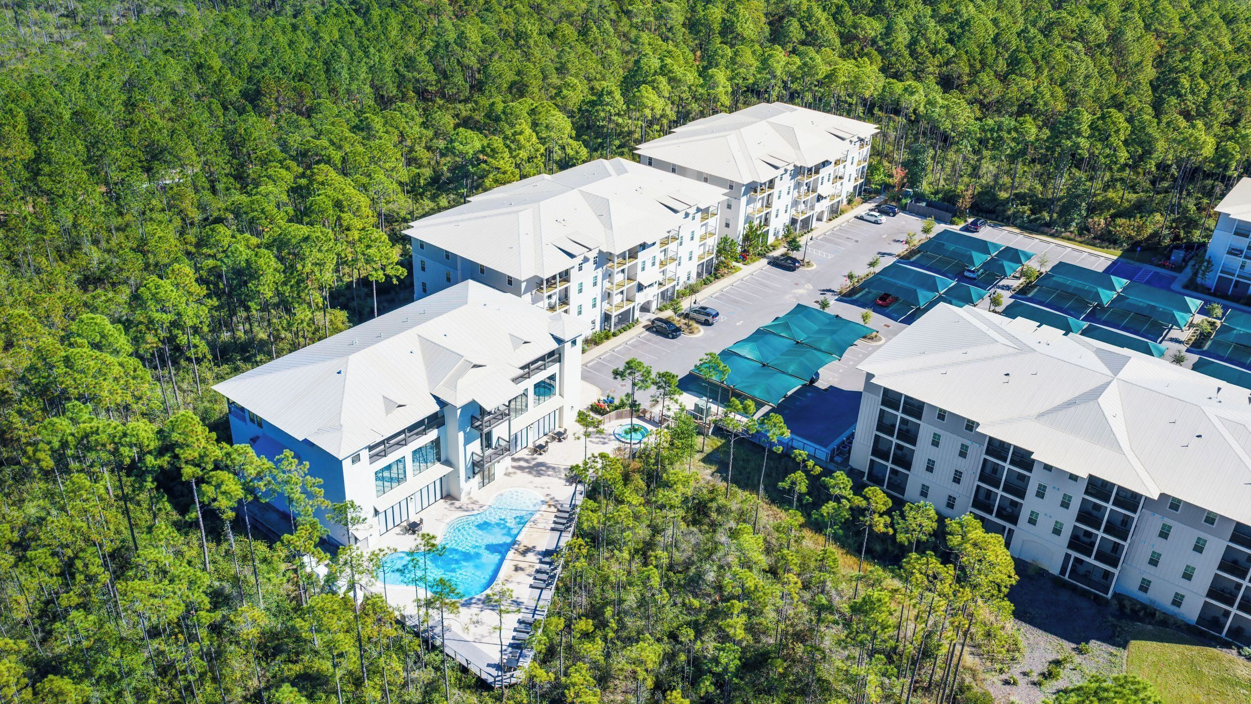 An aerial view of a resort with multiple buildings, a swimming pool, and a parking area surrounded by dense forest.