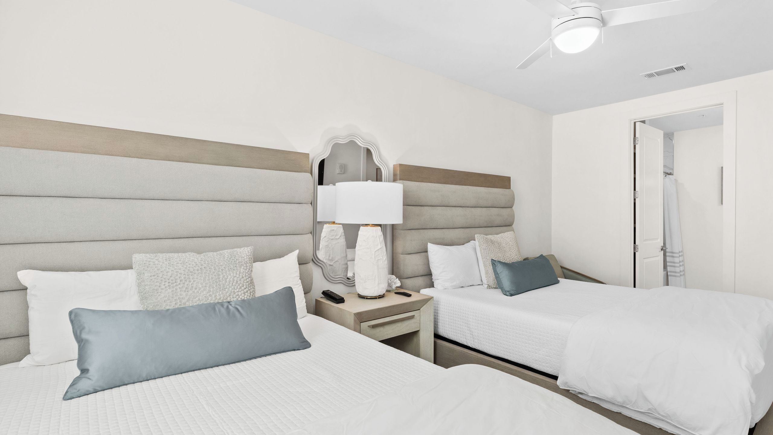 A hotel room with two beds, a nightstand with a lamp, a ceiling fan, and a door leading to a bathroom.