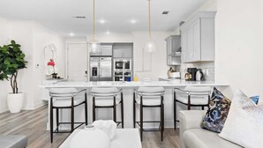 A modern kitchen with a central island, bar stools, and a dining area with a sofa and cushions. - Bright 30A Two-suite Condo Balcony Views, Pool, Hot Tub, Near Gulf Place Emerald Oasis by Avantstay (Santa Rosa Beach)