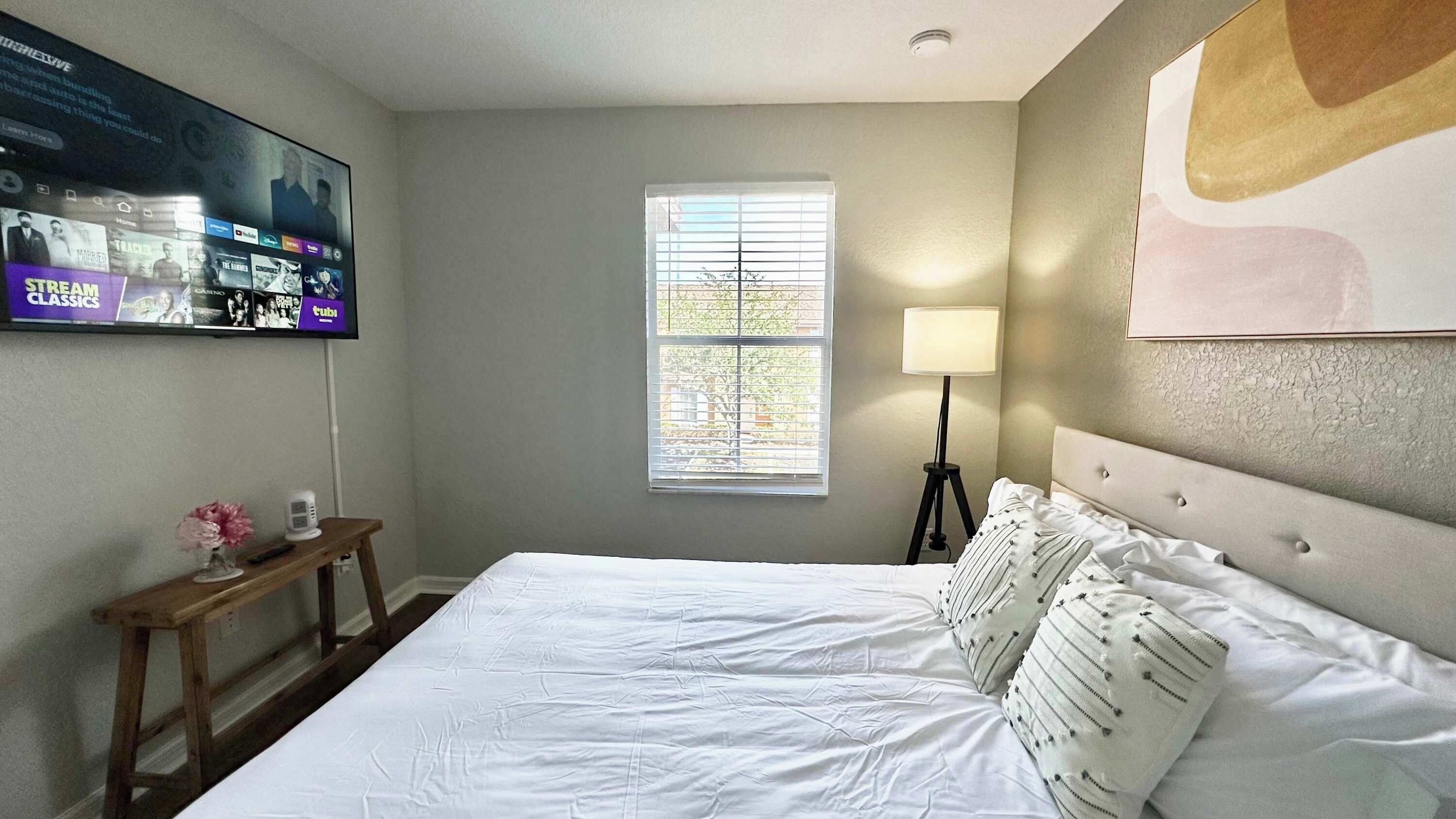 A bedroom with a bed, a TV mounted on the wall, a window with blinds, a bedside lamp, and a small table with a vase.