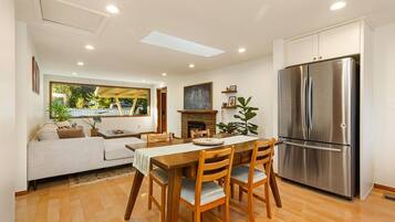 A modern kitchen with a stainless steel refrigerator, wooden dining table, and chairs. There is a fireplace and a view of the outdoors through a large window.