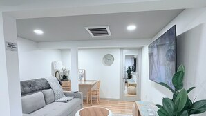 Ett modernt vardagsrum med en grå soffa, en rund träpall, en matplats med ett träbord och stolar samt en stor krukväxt.