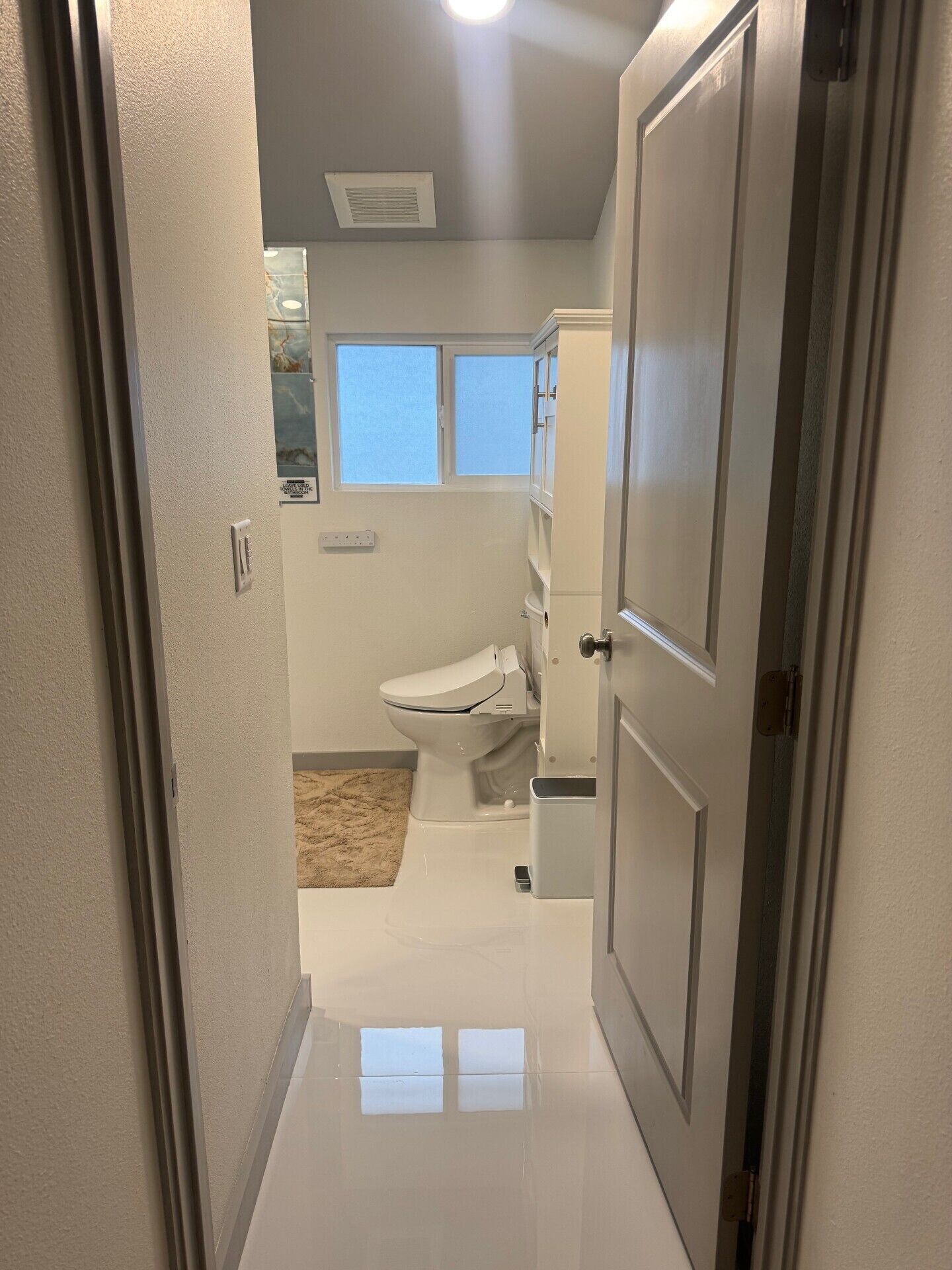 A narrow hallway leading to a bathroom with a toilet, a small window, and a door.