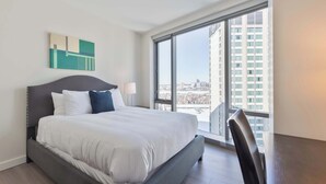 A modern hotel room with a large bed, a view of the city through a floor-to-ceiling window, and a minimalist decor. - Luxury Apt Back Bay | Near Prudential Center (Boston, MA)