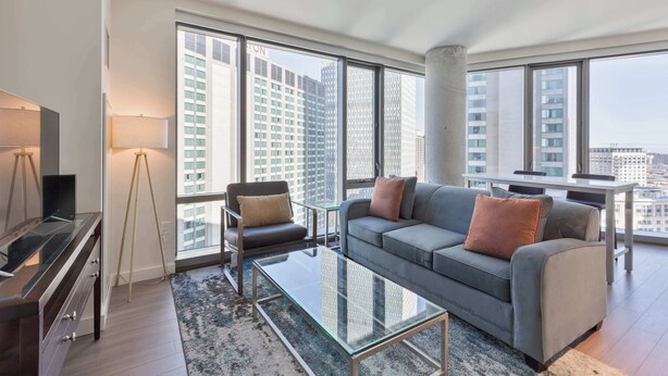 A modern living room with a sofa, armchair, and coffee table, large windows with a city view, and a glass cabinet. - Luxury Apt Back Bay | Near Prudential Center (Boston, MA)
