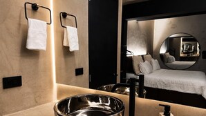 A modern bathroom with a large mirror, a round sink, and a bed in the background.