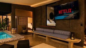 A modern hotel room with a large TV screen displaying 'NETFLIX,' a pool area, a sofa, and a small table with a lamp.