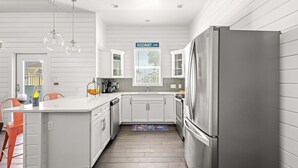 A modern kitchen with white cabinets, a stainless steel refrigerator, and a window with a view of the outside. - Hemingway House, Resort Pools & Pirate Playground (Port Aransas)