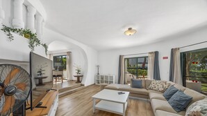 Ein geräumiges Wohnzimmer mit einem weißen Sofa, einem Couchtisch und einem Ventilator. Ein großes Fenster mit Vorhängen bietet Blick auf Grünflächen.
