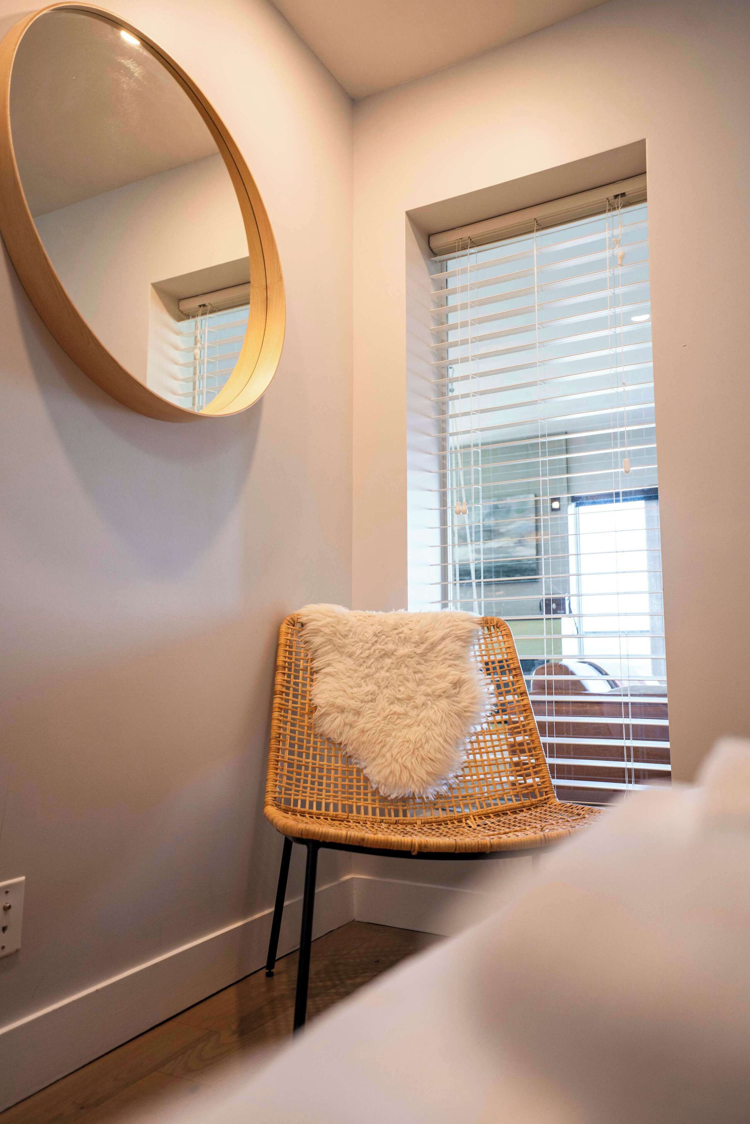 Una habitación con una silla de mimbre, un espejo redondo y una ventana con persianas.