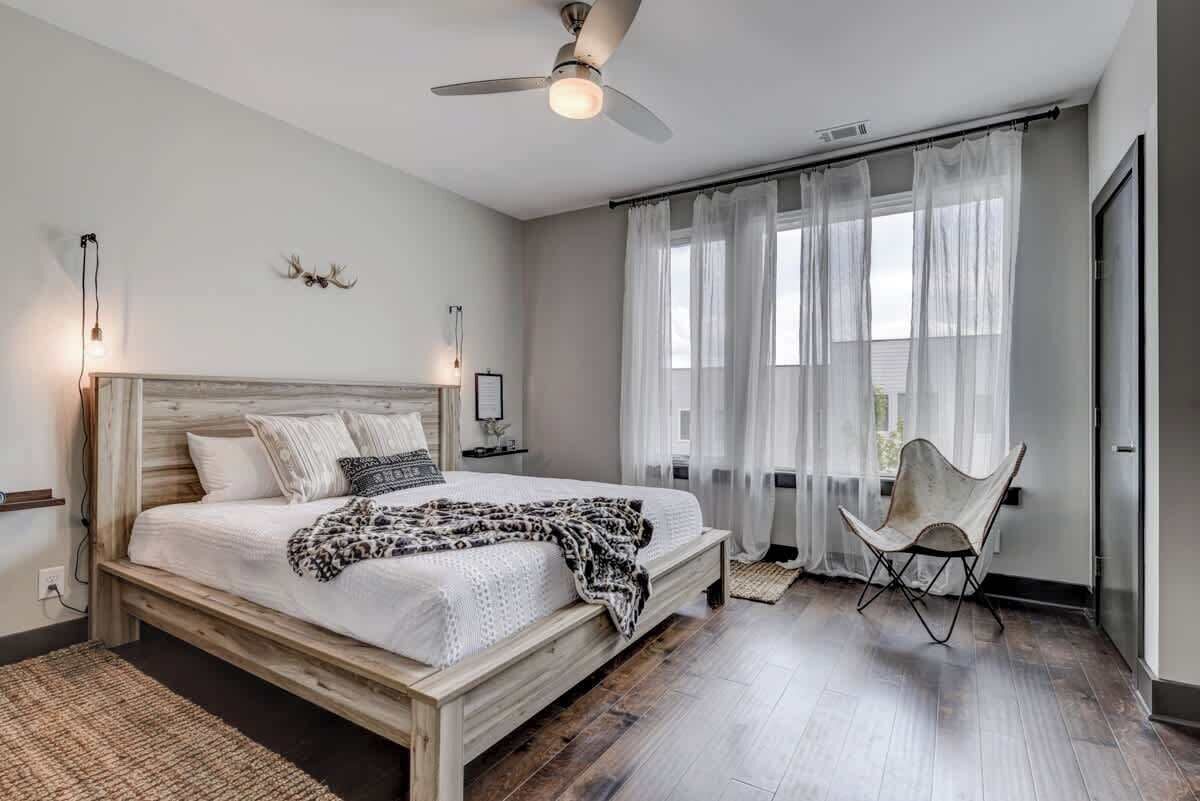 A bedroom with a wooden bed, a ceiling fan, a chair, and a window with sheer curtains.
