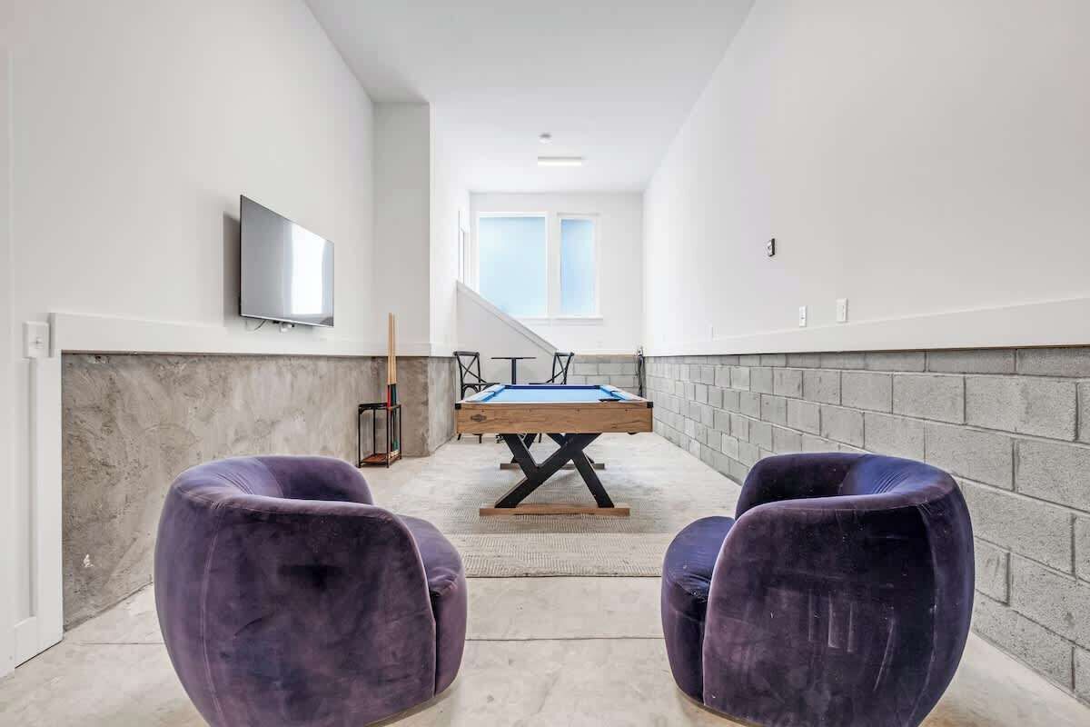 Una habitación moderna con mesa de billar, dos sillas moradas, televisor y una escalera que lleva a un balcón.
