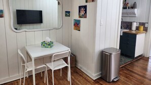 Una habitación con una mesa blanca, dos sillas blancas, un televisor, un bote de basura y una zona de cocina con armarios y un lavamanos.