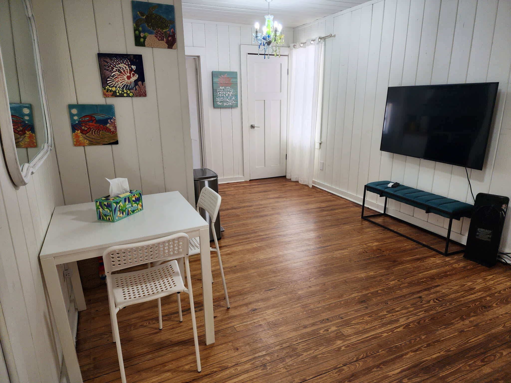 Une pièce avec un plancher en bois, une table blanche avec une boîte de mouchoirs, une chaise blanche, un banc, un téléviseur et des œuvres d’art murales.