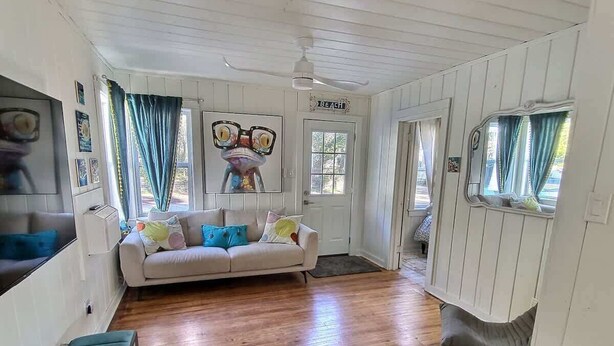 Una habitación con piso de madera, un sofá blanco con cojines de colores, un banco y un televisor montado en la pared.