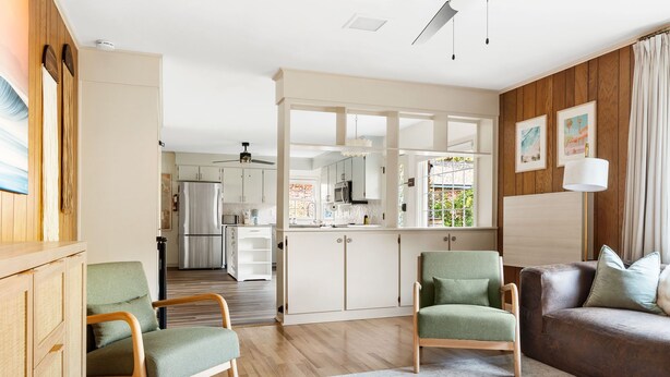 Uma sala de estar moderna com piso de madeira, poltronas verdes, um sofá de couro marrom e uma cozinha ao fundo.