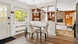 Cozinha moderna com armários brancos, uma área de jantar com mesa e cadeiras brancas, e vista para o exterior por grandes janelas.