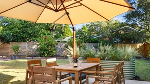 Mesa de madeira no pátio com cadeiras, um grande guarda-sol e uma garrafa de vinho.