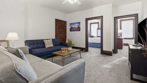 Un salón con un sofá azul, un sofá gris, una mesa de centro de madera y un ventilador de techo. Hay dos puertas que dan a otras habitaciones.