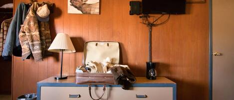 A room with a wooden wall, a coat rack with jackets, a television mounted on the wall, a suitcase on a dresser, and a trash bin.