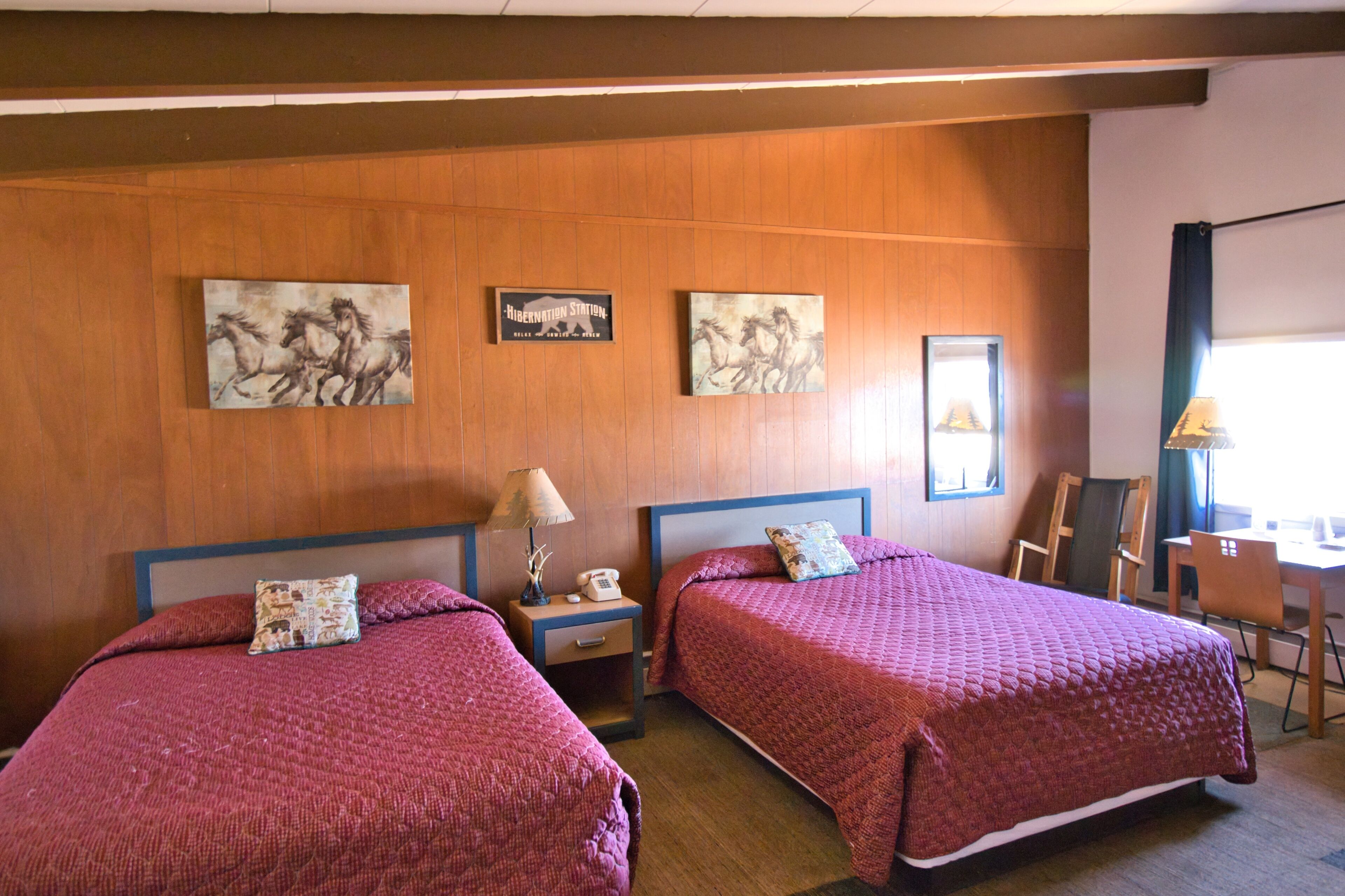 A hotel room with two beds, wooden panel walls, artwork, a desk, and a chair.