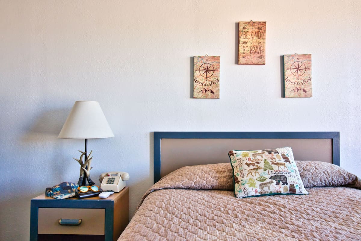 A bedroom with a bed, a nightstand with a lamp, a phone, and a drawer. There are three wall decorations above the bed.